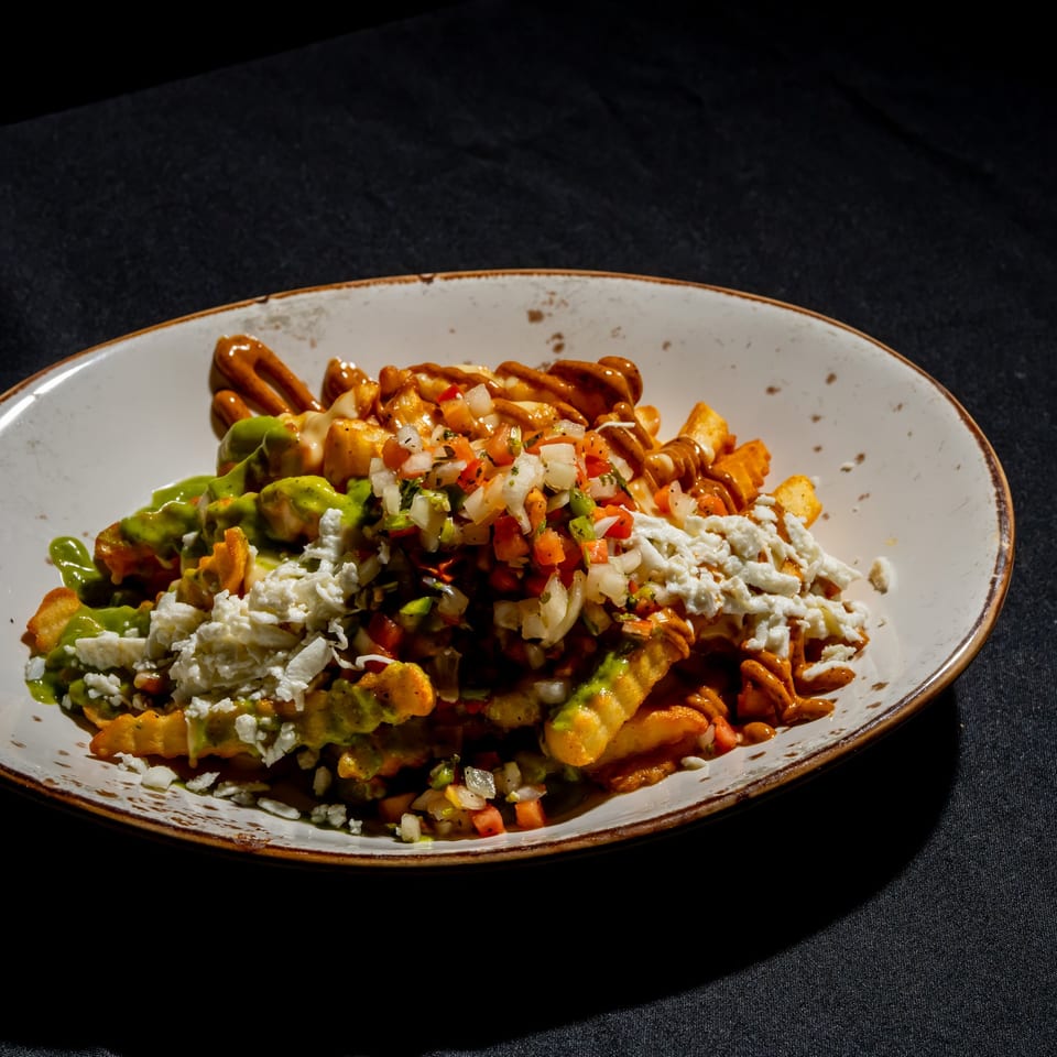 Loaded fries topped with avocado sauce, crumbled cheese, diced vegetables, and drizzled sauces on a ceramic plate.