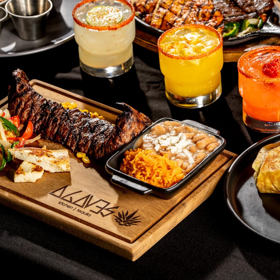 Mexican restaurant spread featuring grilled steak, fajita vegetables, rice, beans, quesadillas, tacos, and three chili-rimmed cocktails.