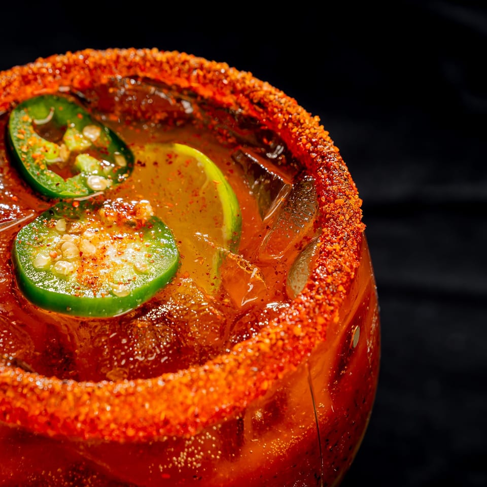 Close-up of a spicy red cocktail in a glass with a chili-coated rim, ice, and lime slices against a black background.