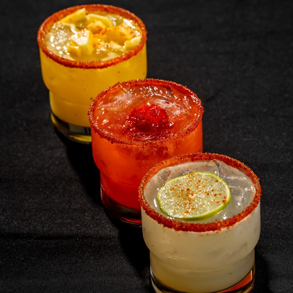 Three colorful cocktails with chili-salt rims, ice, and citrus or fruit garnishes arranged on black fabric.