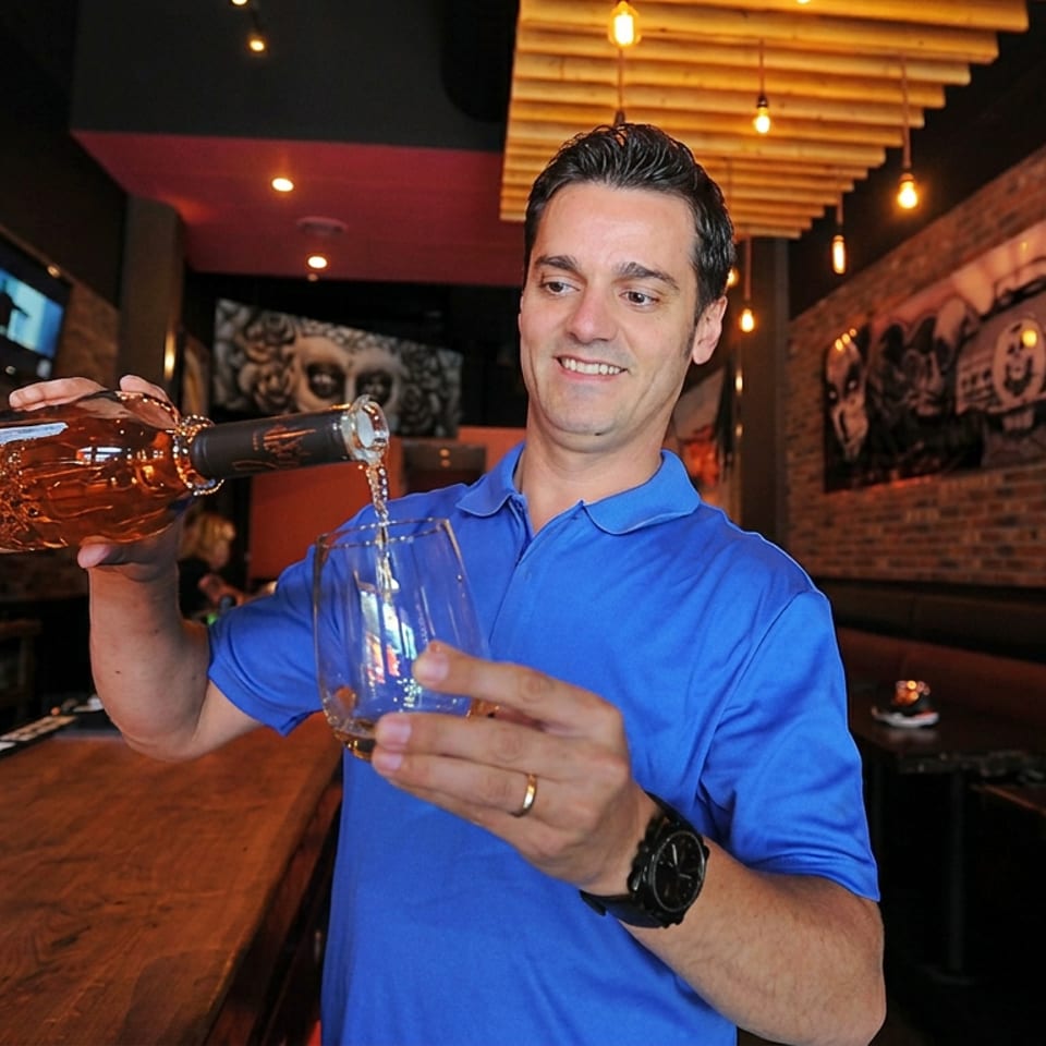 Bartender pours amber liquor from a bottle into a glass inside a warmly lit, brick-walled restaurant bar.