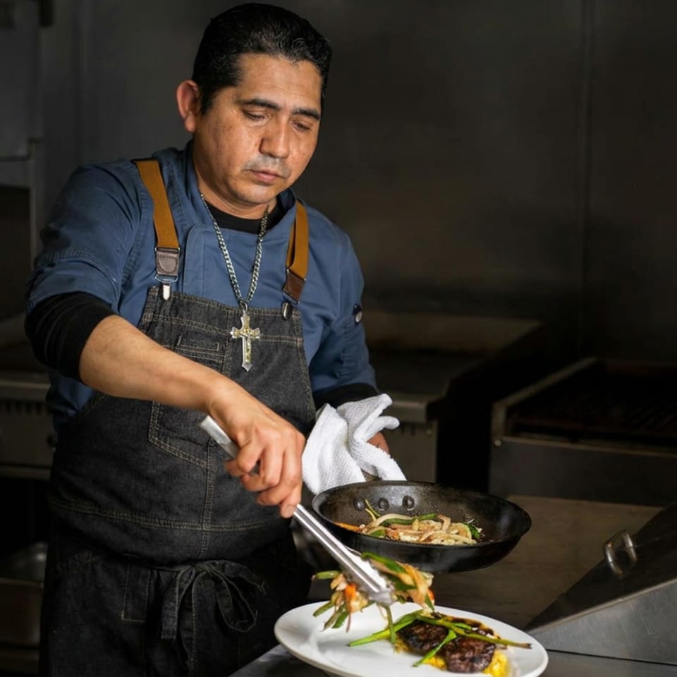 Chef in a commercial kitchen plates grilled vegetables and meat from a skillet onto a white dish.