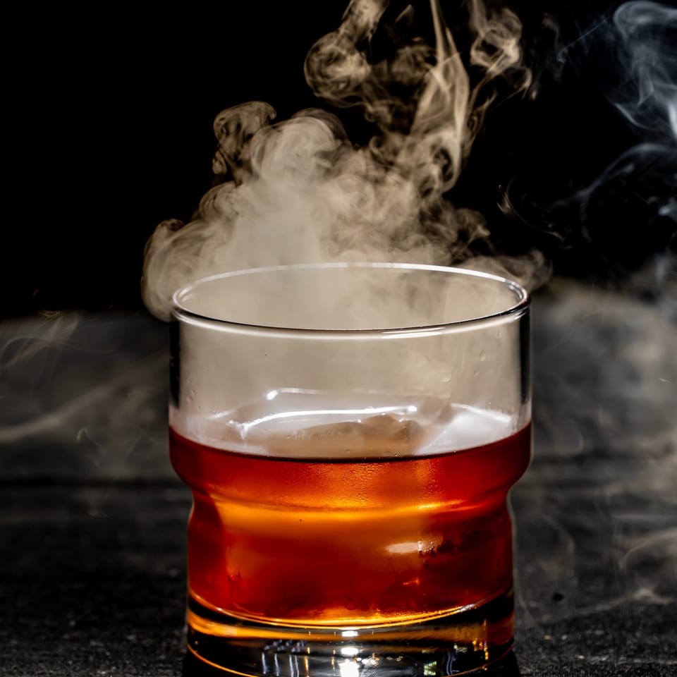 Amber cocktail in a clear rocks glass emits dramatic swirling smoke against a dark background.