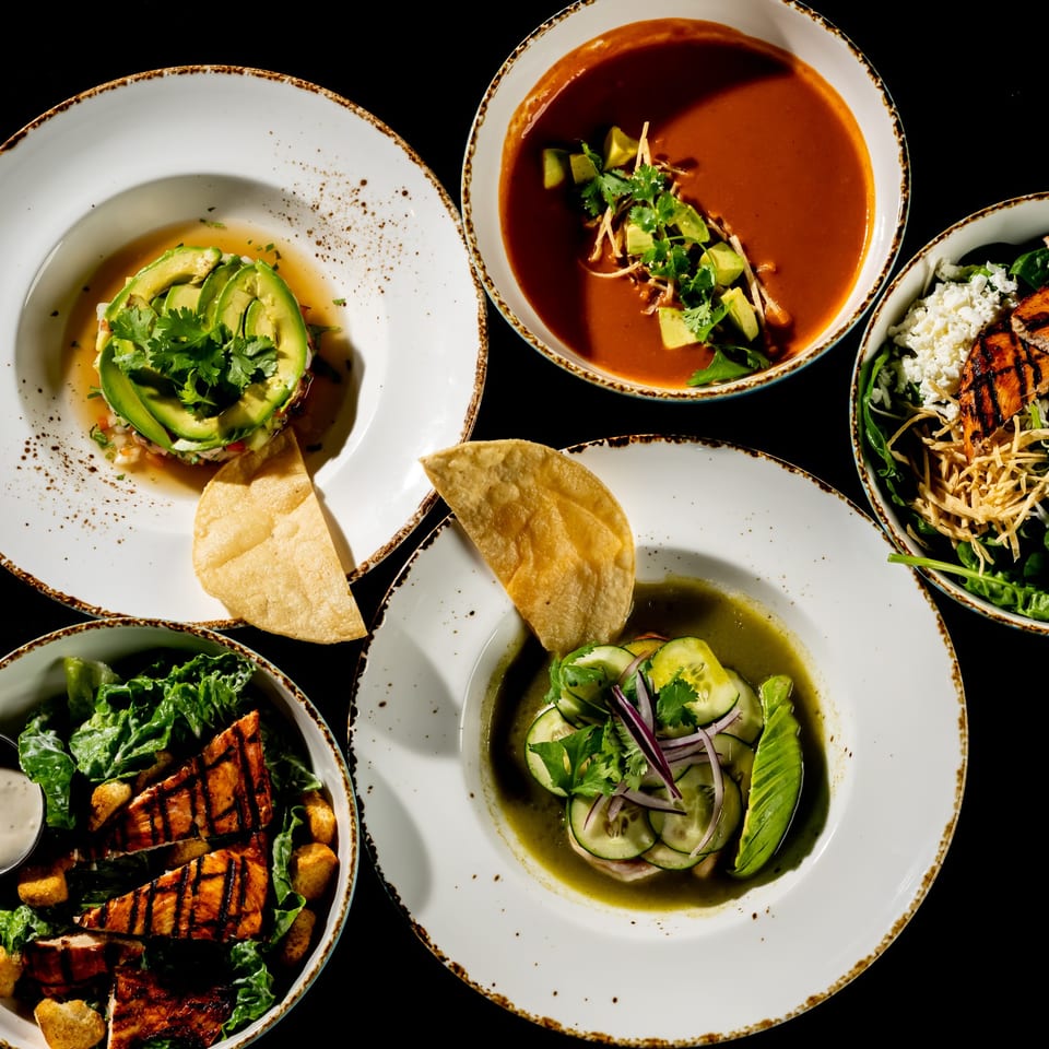 Overhead spread of colorful restaurant dishes, including grilled salmon salads, avocado-topped entrée, soup, and vegetable plates.