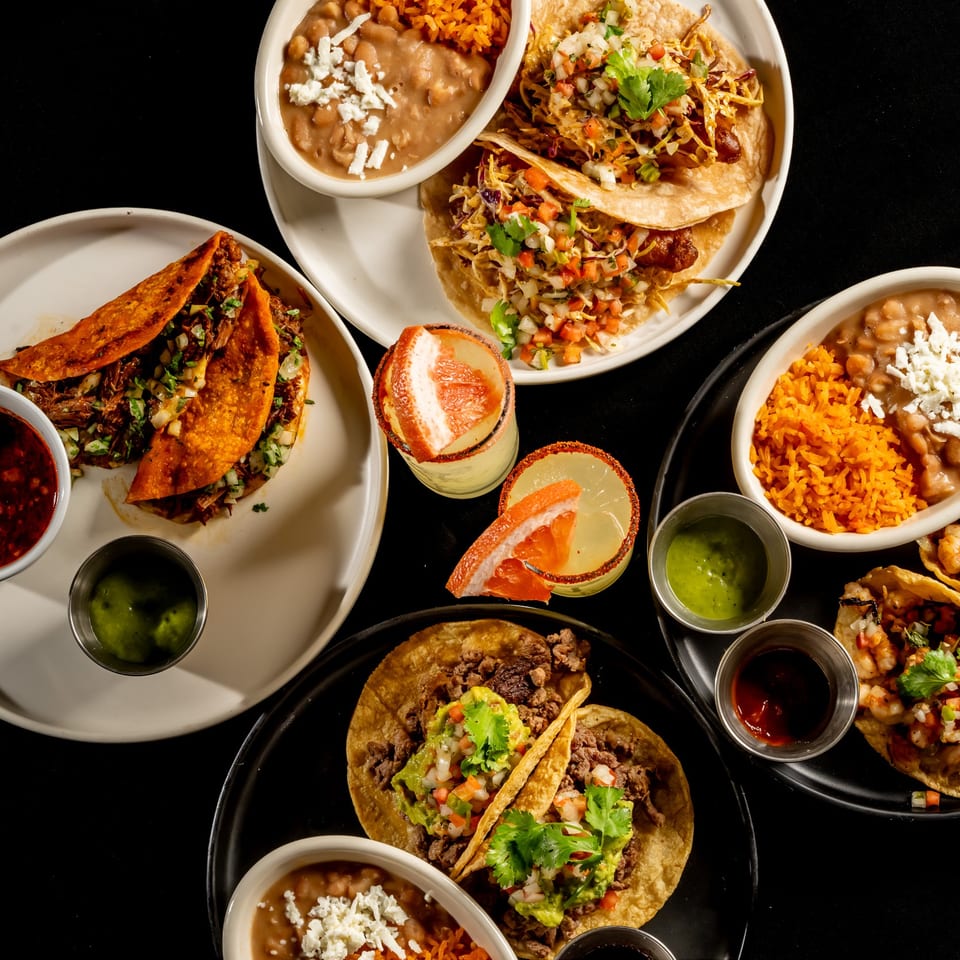 Overhead spread of Mexican tacos, quesadillas, rice, beans, salsas, garnished tostadas, and citrus cocktails on black tables.