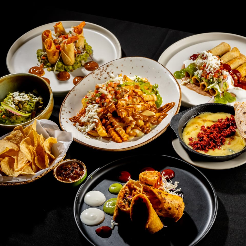 Assorted Mexican-inspired dishes, including taquitos, saucy pasta, guacamole, chips, nachos, and cheesy dip, arranged on a black table.