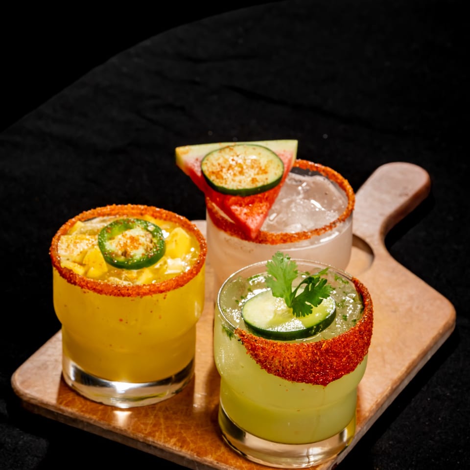 Three colorful cocktails with chili-salt rims, cucumber and jalapeño garnishes, arranged on a wooden serving board.