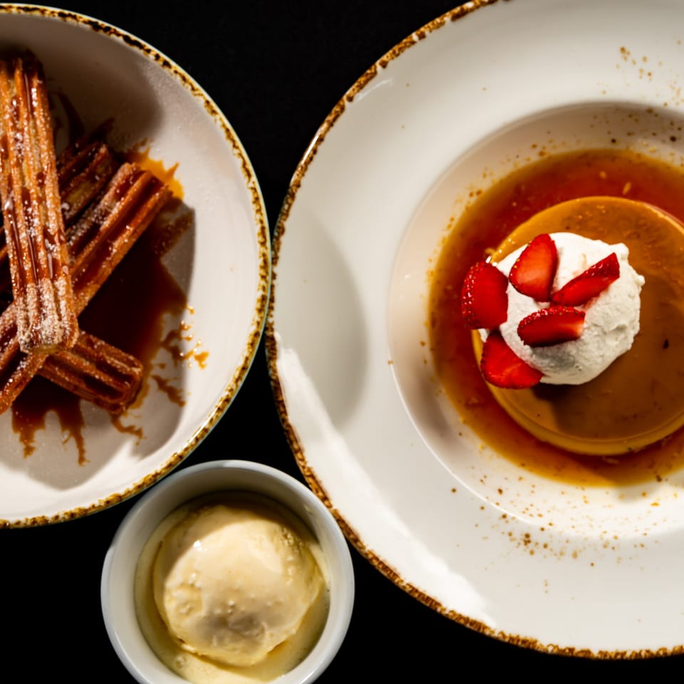 Overhead view of churros with chocolate dipping sauce beside flan topped with whipped cream and strawberries, plus vanilla ice cream.