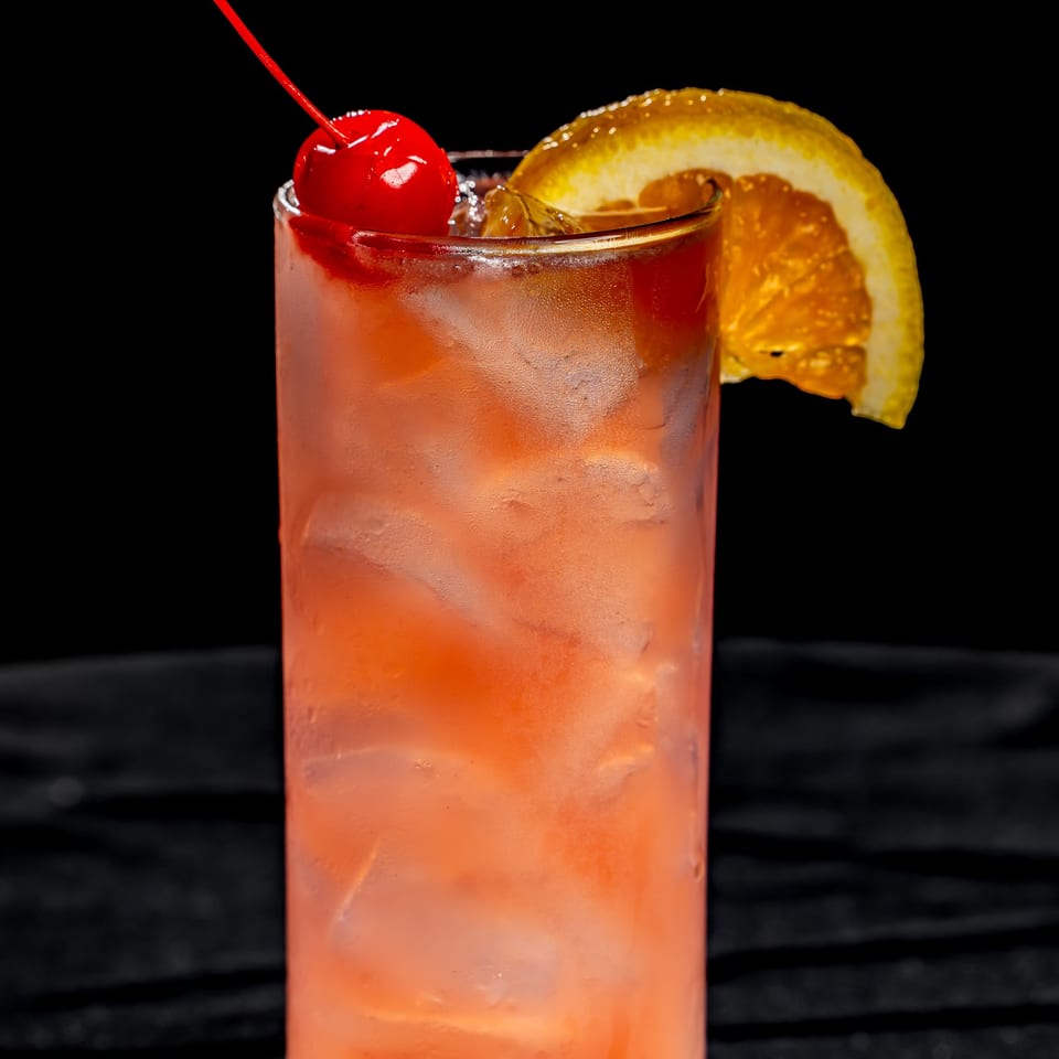 Tall chilled pink cocktail garnished with a maraschino cherry and orange slice, photographed against a black background.