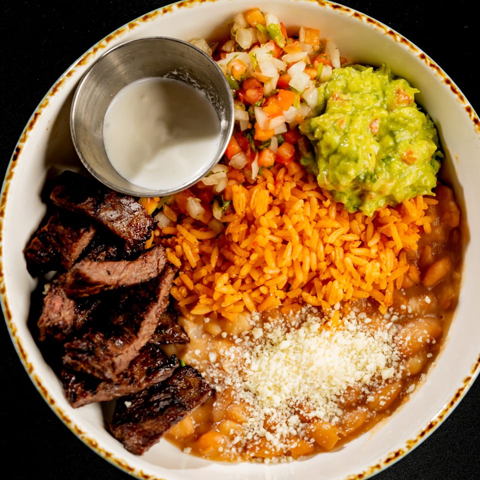 Overhead view of a Mexican-inspired plate with grilled steak, orange rice, beans topped with cheese, guacamole, salsa, and crema.