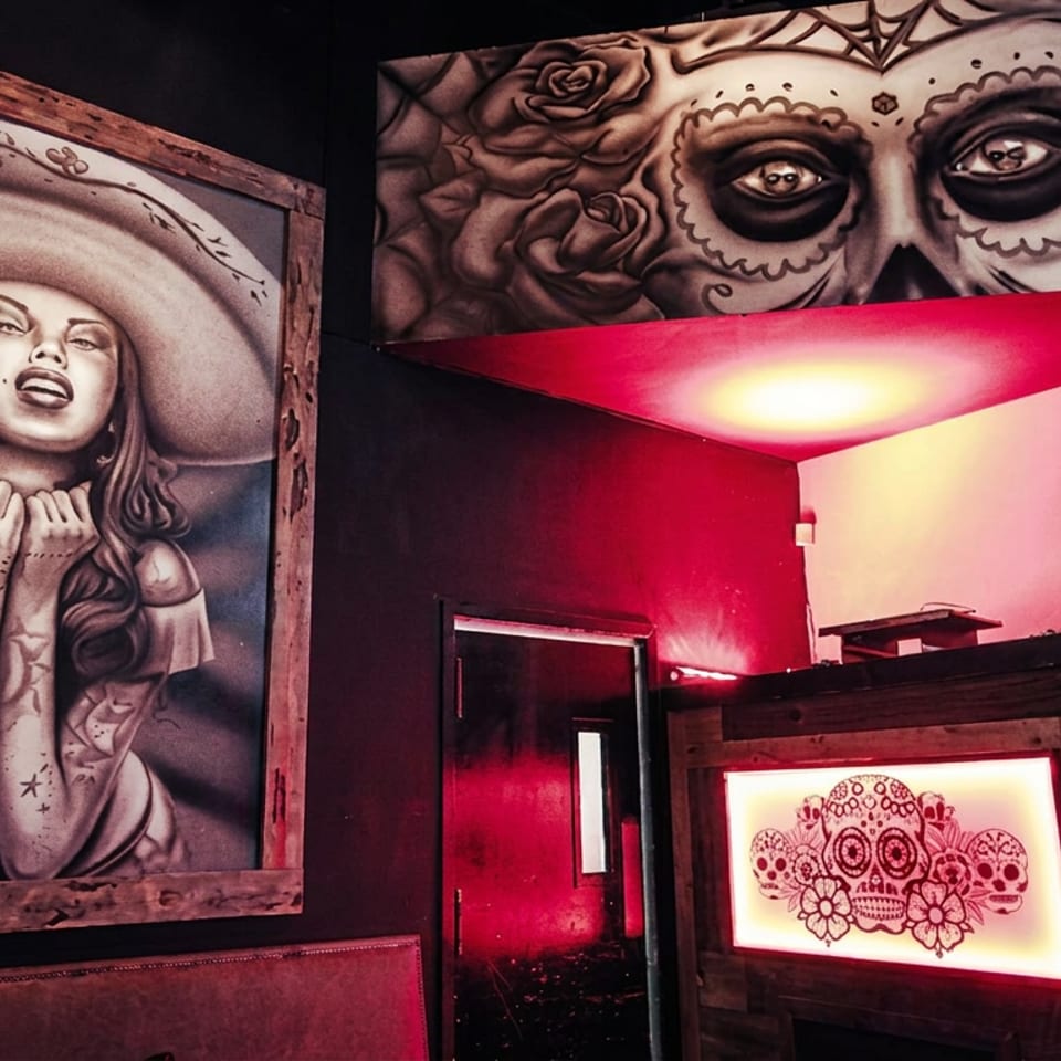 Dark restaurant interior featuring Mexican-inspired murals of a woman, skulls, and roses, illuminated by dramatic red lighting.
