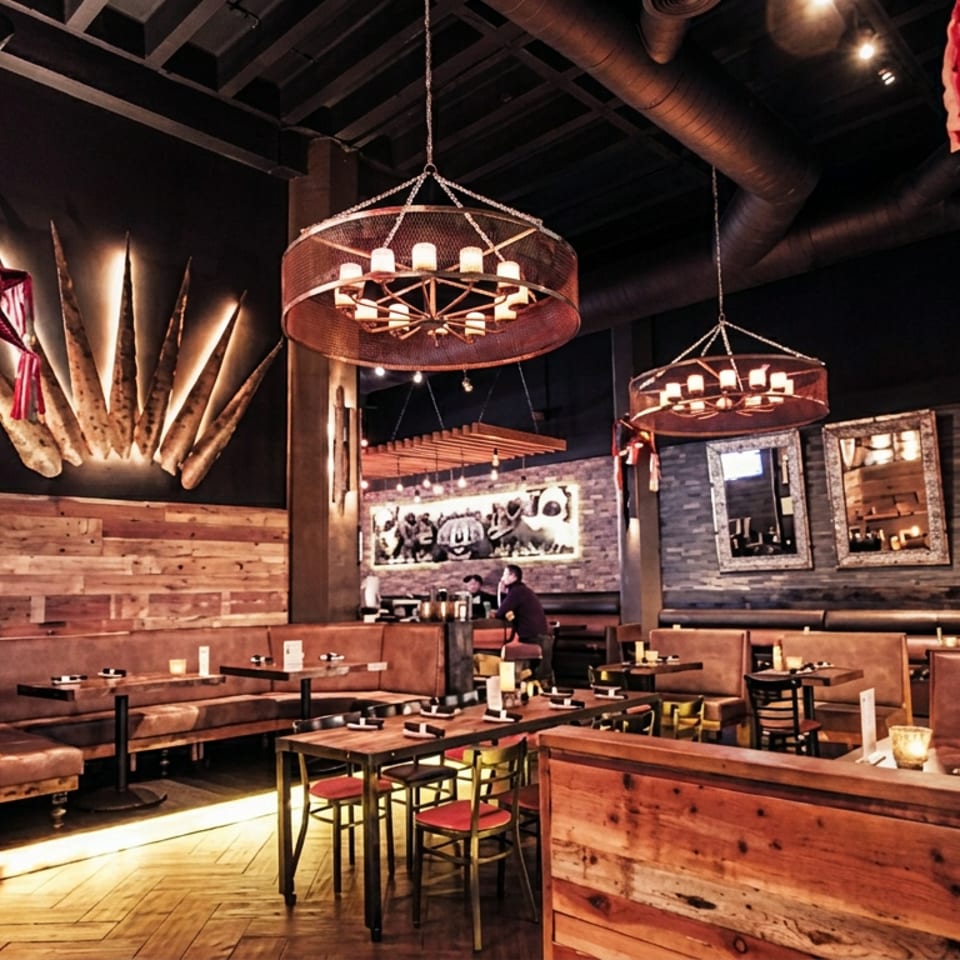 Warmly lit restaurant interior with wood-paneled walls, leather booths, set dining tables, hanging circular chandeliers, and rustic decor.