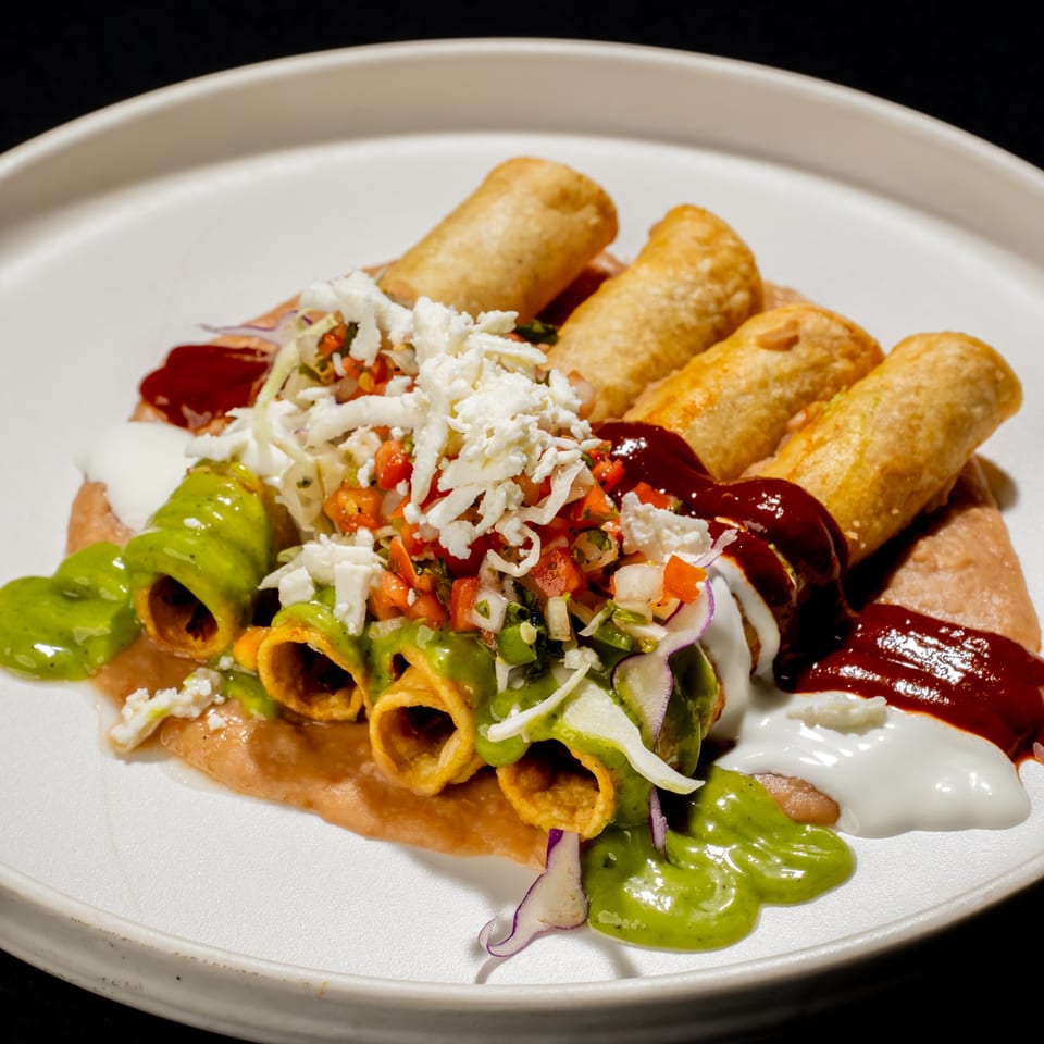 Crispy rolled tacos topped with lettuce, pico de gallo, crumbled cheese, green sauce, red sauce, and sour cream on a white plate.