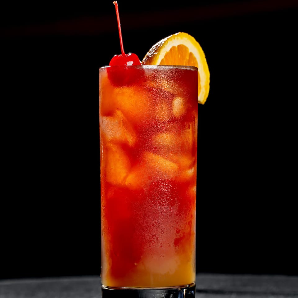 Iced red-orange cocktail in a tall glass, garnished with a cherry and orange slice against a dark background.