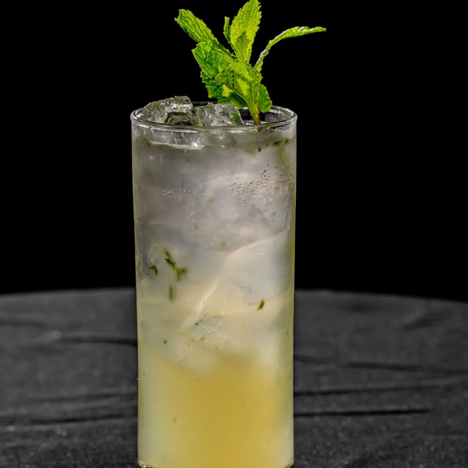 Tall glass of pale yellow mint cocktail with ice, condensation, and fresh mint garnish on a dark table.